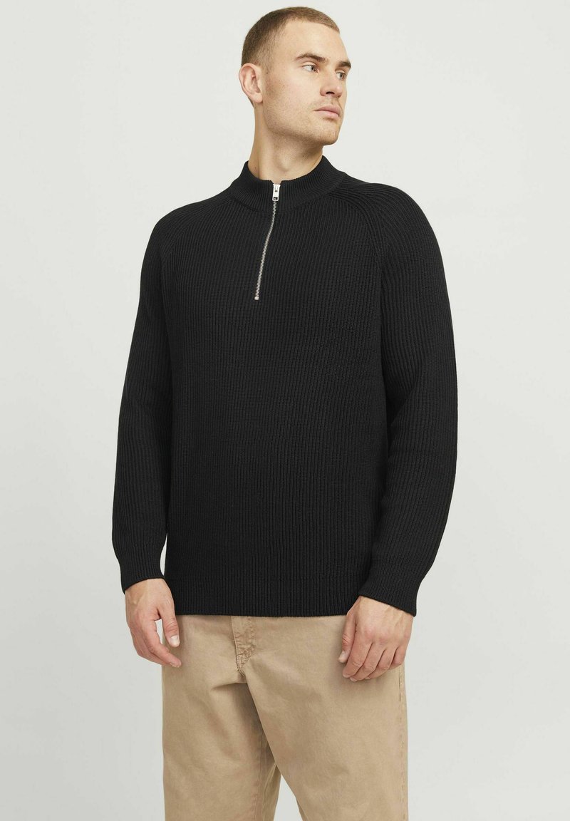 Schwarzer Rippstrick-Pullover mit hohem Kragen und halblanger Reißverschluss. Verfügt über lange Ärmel und eine lockere Passform. Getragen mit beigen Hosen.