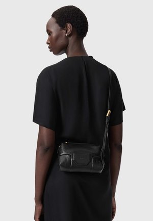 Frau mit kurzen Haaren, die ein schwarzes Kleid trägt und eine schwarze Leder-Umhängetasche über der Schulter vor einem einfarbigen Hintergrund trägt.