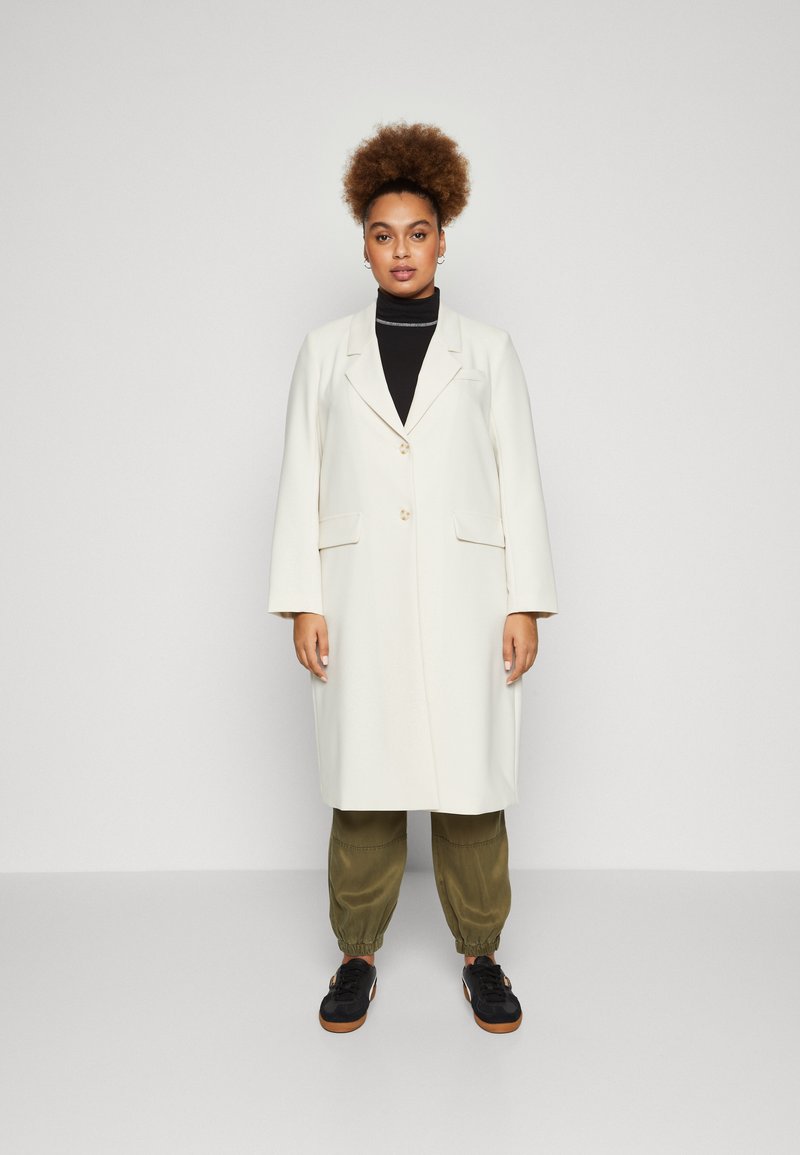 Lang hvid frakke med notched lapel, to knapper og sidelommer, båret over en sort rullekrave og olivengrønne bukser. Sorte sneakers.