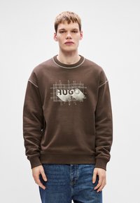 Brun bomullssweatshirt med ribbad halsringning och muddar, med en ljusfärgad grafisk design som inkluderar texten "HUGO." Avslappnad passform.