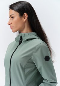 Lys grøn hættejakke lavet af glat stof, med en central lynlås og et struktureret sort logo patch på venstre ærme.