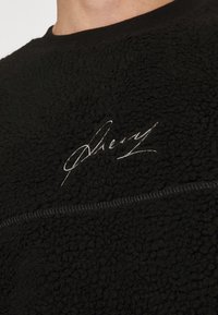 Pullover in fleece nero con superficie testurizzata, dettaglio firmato ricamato bianco e cucitura a contrasto lungo la cucitura superiore.