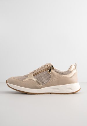Baskets de sport beiges avec des panneaux en maille, des accents en cuir lisse, un détail zippé sur le côté et une semelle blanche rembourrée.