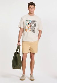 Beeži värvusega graafiline t-särk värvilise trükiga, helerohelised lühikesed püksid, oliivrohelise värvusega kandekott ja helepruunid tossud, seisab neutraalse tausta ees.