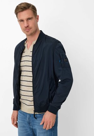 Mand iført navy bomberjakke over beige stribet sweater og blå jeans mod en ensartet hvid baggrund.