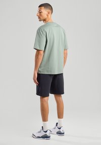 Light green cotton t-shirt, short sleeves, round neckline, paired with dark shorts. Model stands with back to camera, wearing white sneakers.