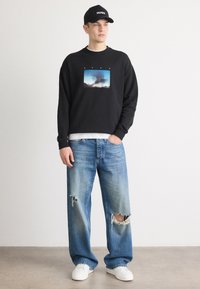 Felpa nera con girocollo e stampa grafica, abbinata a jeans chiari strappati e sneakers bianche, completata da un cappellino nero.