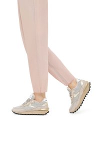 Lichtgrijze en beige sneakers met metalen gouden accenten, nubuck en gestructureerde stof, en een dikke zwarte zool. Gestyld met roze tapered broeken.