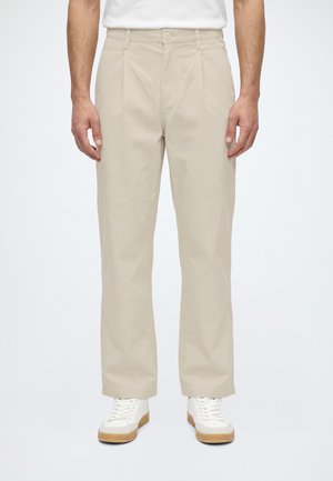 Homme portant un pantalon beige droit avec des plis, des baskets blanches à semelles en gomme, et un t-shirt blanc rentré.