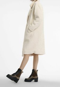 Kremowy płaszcz z futrzanym sztucznym futerkiem, z długimi rękawami, bocznymi kieszeniami i kołnierzem; zestawiony z brązowymi i czarnymi sznurowanymi botkami na obcasie.