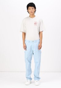 Camiseta de manga corta gris claro con diseño gráfico rojo, combinada con pantalones cargo azul claro. El modelo se encuentra con una postura neutral frente a un fondo liso.