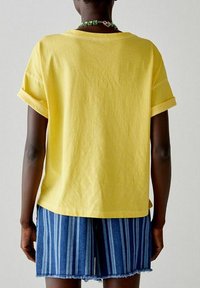 T-shirt di cotone giallo con maniche arrotolate, abbinato a pantaloni corti in denim blu con strisce verticali. Design semplice, vestibilità rilassata.
