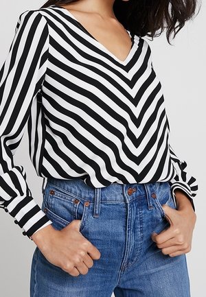 Femme portant un chemisier à manches longues à rayures chevron noires et blanches, rentré dans un jean bleu taille haute, les mains dans les poches.