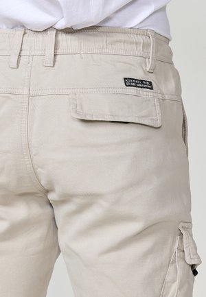 Beige Cargo-Hosen mit einer strukturierten Oberfläche, ausgestattet mit einer Gesäßtasche und einem Etikett. Das Design umfasst Gürtelschlaufen und eine Seitentasche.