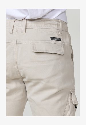 Beige Cargo-Hosen mit einer strukturierten Oberfläche, ausgestattet mit einer Gesäßtasche und einem Etikett. Das Design umfasst Gürtelschlaufen und eine Seitentasche.