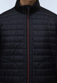 Personne portant une veste matelassée noire avec un col montant et une fermeture éclair visible sur le devant de couleur rouge foncé contrastante.