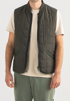 Man draagt een beige shirt met korte mouwen, een groen doorgestikt mouwloos vest met een rits aan de voorkant, en een groene broek tegen een effen lichte achtergrond.