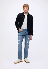 Ung mand stående med hånd i lommen iført sort skjorte med knapper, hvid T-shirt, revnede lyseblå jeans og hvide sneakers.