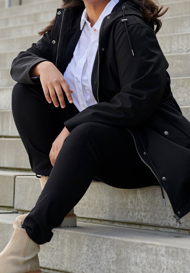 Schwarze Jacke mit Reißverschluss und Kapuze über einem weißen Hemd, kombiniert mit schwarzen Hosen und beigen Stiefeletten, sitzend auf grauen Stufen.