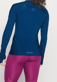 Granatowa sportowa bluza z długim rękawem z fakturowanymi akcentami na ramionach, tkaniną odprowadzającą wilgoć oraz logo DRI-FIT na dolnej części pleców, w połączeniu z różowymi legginsami.