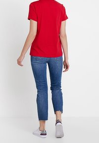Una persona está de pie con la espalda hacia la cámara, vistiendo una camisa roja, jeans azules y zapatillas blancas, contra un fondo blanco liso.