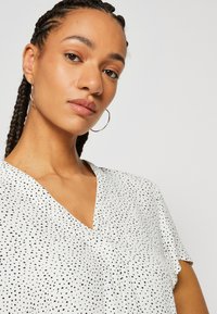 Blusa a V in bianco con pois neri. Tessuto leggero con maniche corte e vestibilità ampia. Presenta una sottile piega al centro.