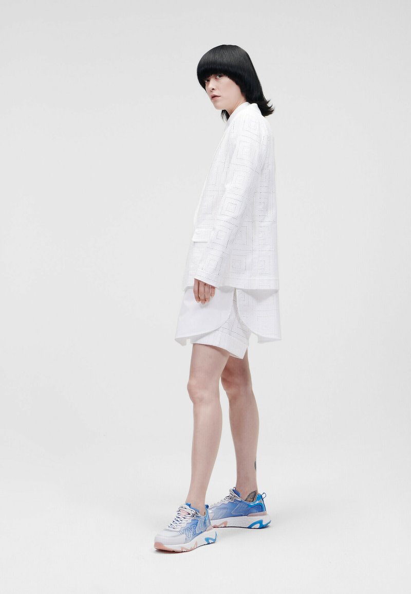 Weißes, strukturiertes Übergrößenhemd mit geometrischem Muster, kombiniert mit weißen Shorts. Die Schuhe haben blaue Akzente und ein mehrfarbiges Design.