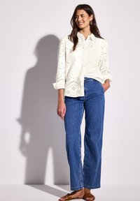 Valkoinen nappipaita, jossa on koristeellisia silmäleikkauksia, yhdistettynä sinisiin korkeavyötäröisiin farkkuihin ja ruskeisiin litteisiin sandaaleihin.