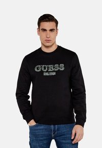 Felpa nera con collo tondo, caratterizzata dal testo "GUESS EST. 1981" in verde a contrasto, con polsini e orlo a coste, indossata sopra jeans blu.