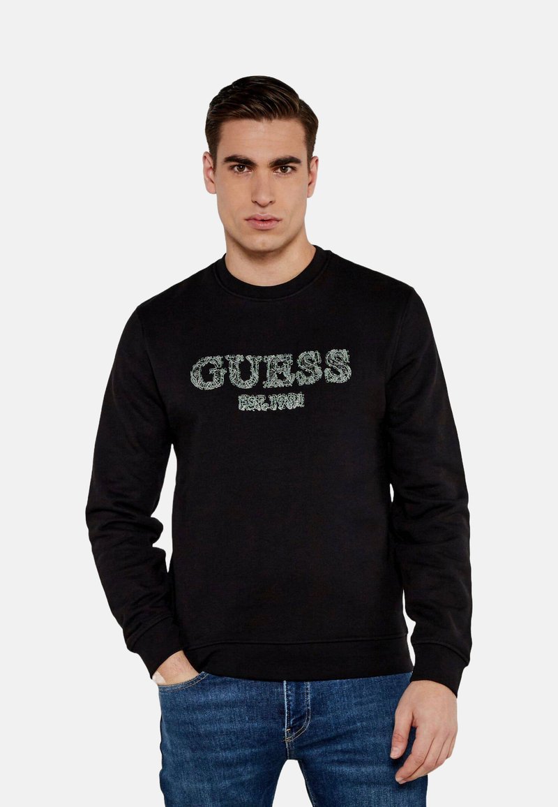 Felpa nera con collo tondo, caratterizzata dal testo "GUESS EST. 1981" in verde a contrasto, con polsini e orlo a coste, indossata sopra jeans blu.