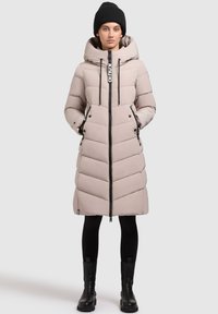 Garšs, gaiši beidž krāsas pūkains mētelis ar kapuci, ar ševronu šuvēm, melnu rāvējslēdzēju un akcentiem, valkāts virs melnām legingām un zābakiem.