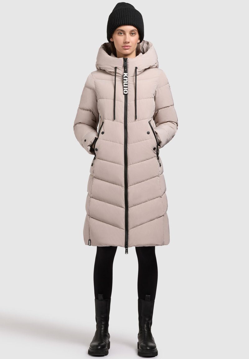 Garšs, gaiši beidž krāsas pūkains mētelis ar kapuci, ar ševronu šuvēm, melnu rāvējslēdzēju un akcentiem, valkāts virs melnām legingām un zābakiem.