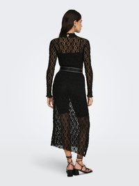 El vestido de encaje negro presenta mangas largas, un cuello alto y una cintura ajustada con una falda transparente. El diseño incluye un borde con forma de ondas y una espalda descubierta.