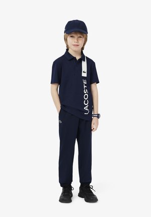Navy polo shirt with white Lacoste logo, paired with matching navy pants and a cap. Black sneakers complete the outfit.