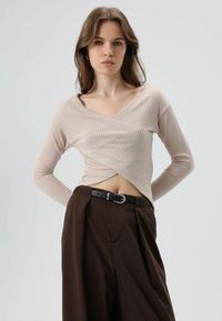 Beige geribd shirt met lange mouwen, diepe V-hals en overslagontwerp, gecombineerd met hoge taille bruine broek en een zwarte riem.