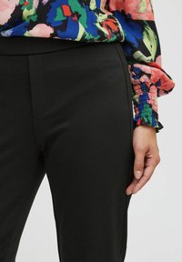 Woman wearing black fitted pants and a multicolored floral blouse with smocked cuffs, standing with one hand relaxed by her side.