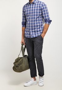 Blau-weiß karierte Hemd mit Knöpfen, graue Slim-Fit-Hosen, weiße Sneakers, trägt eine olivgrüne Canvas-Reisetasche.