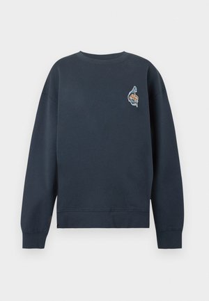 Femme debout, vêtue d'un sweat-shirt bleu marine oversize avec un petit logo sur la poitrine, un jean large bleu clair et des talons ouverts noirs.
