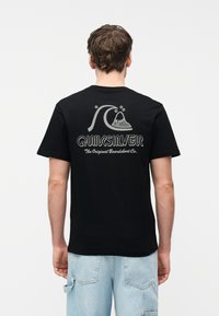 Άνδρας που φοράει μαύρο t-shirt Quiksilver με λογότυπο κύματος και βουνού και κείμενο στην πλάτη, σε συνδυασμό με ανοιχτόχρωμα μπλε τζιν σορτς.