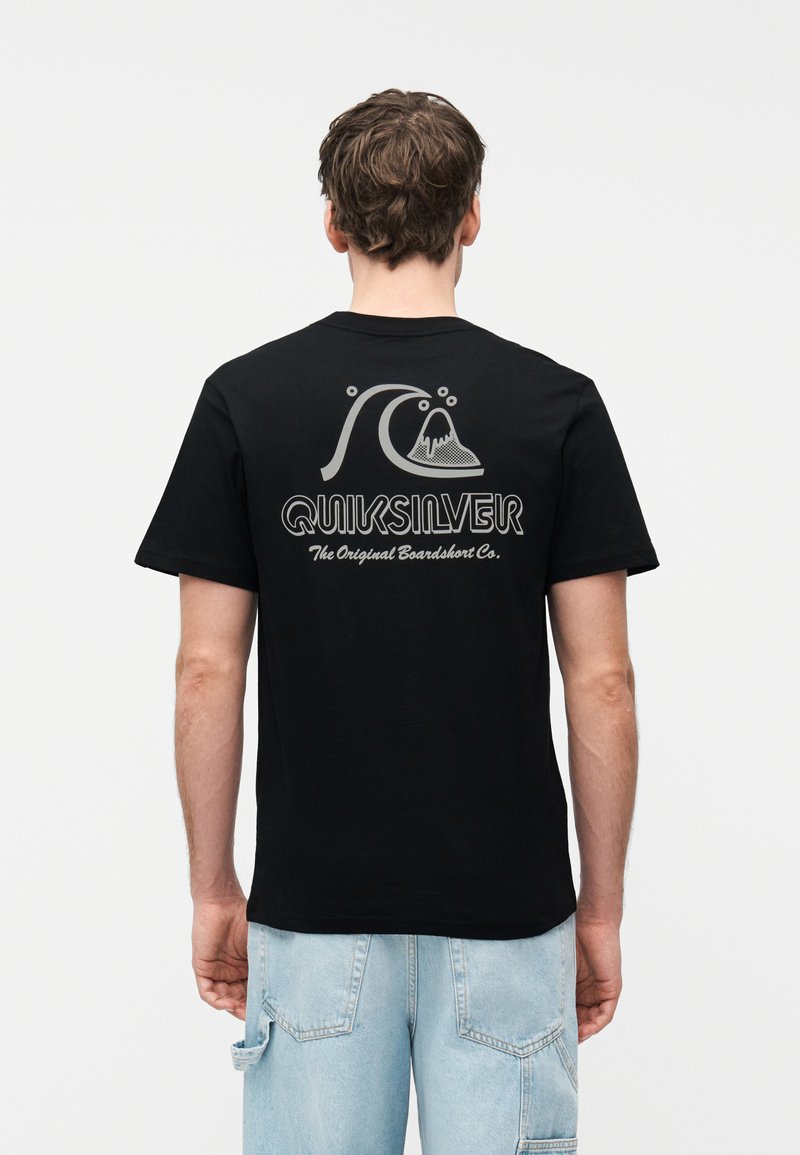 Άνδρας που φοράει μαύρο t-shirt Quiksilver με λογότυπο κύματος και βουνού και κείμενο στην πλάτη, σε συνδυασμό με ανοιχτόχρωμα μπλε τζιν σορτς.