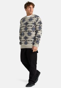 Suéter beige texturizado con patrones geométricos azules y marrones, diseño de cuello redondo, combinado con pantalones negros y zapatos oscuros.