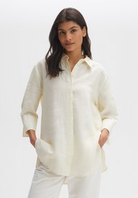 White linen button-up shirt with a relaxed fit, long sleeves, and side pockets. Features a classic collar and light texture.