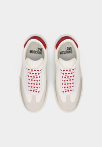 Λευκά sneakers Love Moschino με κορδόνια με κόκκινο μοτίβο καρδιάς και κόκκινες πινελιές στη φτέρνα, εμφανιζόμενα από πάνω σε λευκό φόντο.