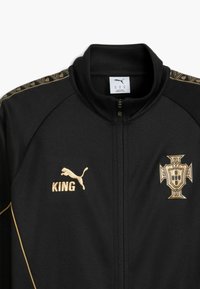 Μαύρο αθλητικό σακάκι με χρυσές λεπτομέρειες. Διαθέτει υψηλό γιακά, φερμουάρ μπροστά, λογότυπο Puma, κείμενο "KING" και εθνικό έμβλημα Πορτογαλίας στο στήθος.