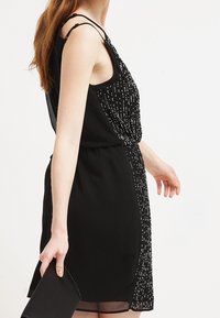 Czarna sukienka na ramiączkach bez rękawów z dopasowanym gorsetem i koralikową detalizacją wzdłuż boku, z cienkimi ramiączkami i przezroczystym dołem.