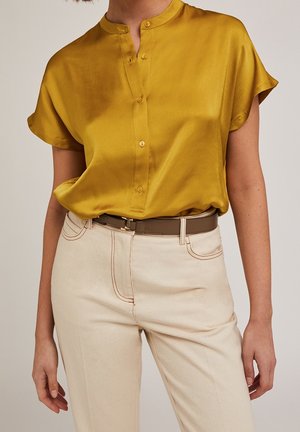 Vrouw draagt een mosterdgele korte mouwen zijden blouse, ingestopt in hooggetailleerde offwhite broek met bruine riem.