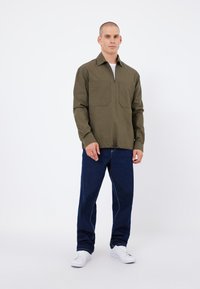 Olivgrünes Langarmhemd mit Halb-Reißverschlusskragen und zwei vorderen Taschen, kombiniert mit blauen Jeans und weißen Turnschuhen.