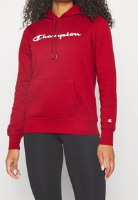 Červená mikina s kapucí a bílým logem "Champion", šňůrková kapuce, přední kapsa, manžety a lem z pružného materiálu. Spárována s černými legínami.
