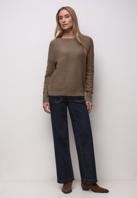 Pull tricot marron clair avec ourlet côtelé, associé à un jean large bleu foncé avec des coutures contrastées. Des bottines marron complètent le look.