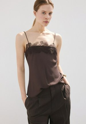 Donna che indossa una canottiera nera con rifiniture in pizzo e pantaloni neri a vita alta, in piedi con le mani nelle tasche su sfondo semplice.